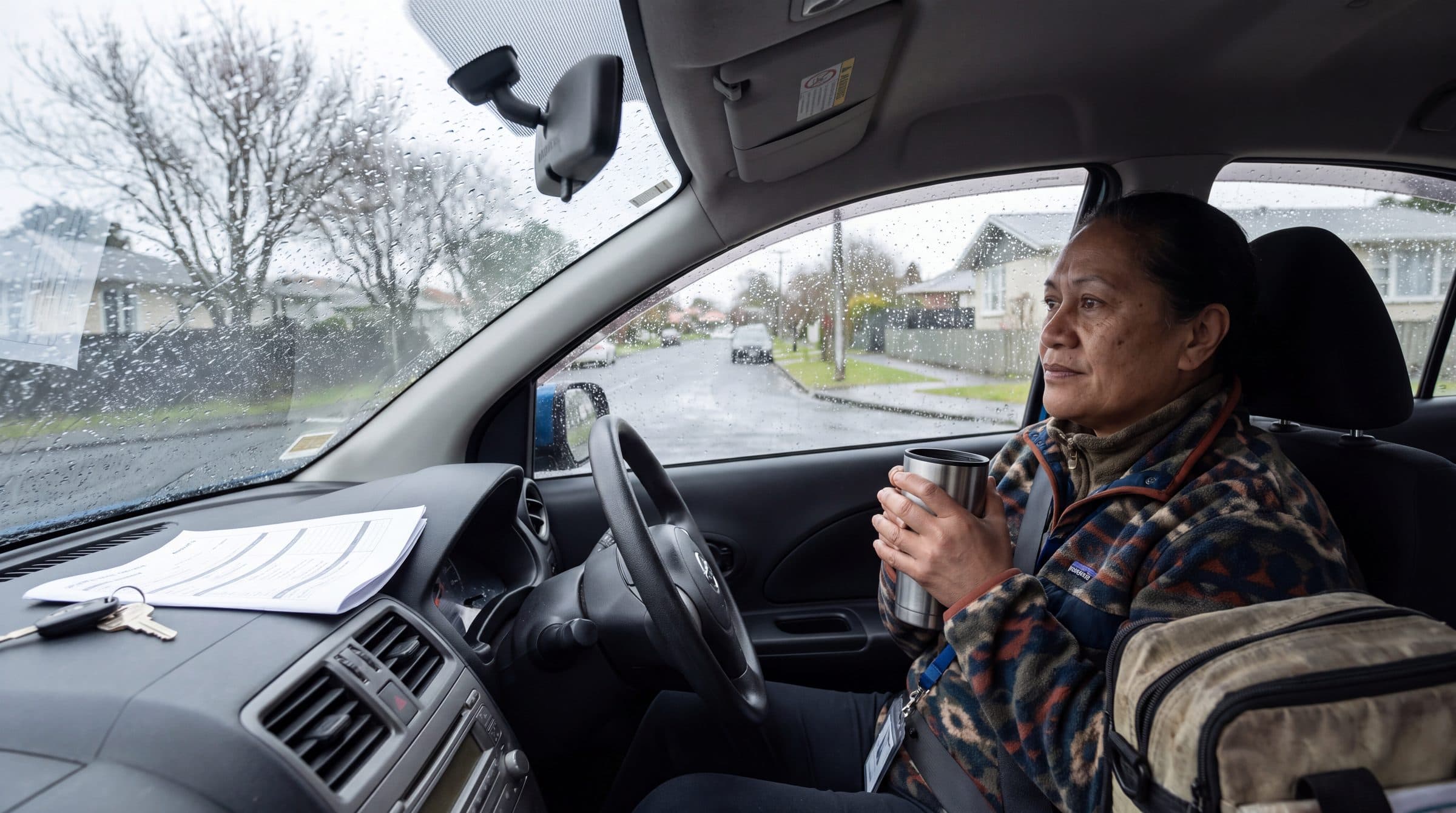A community health worker sits in her car between visits, holding a flask cup, with paperwork and keys on the dashboard and rain on the windows