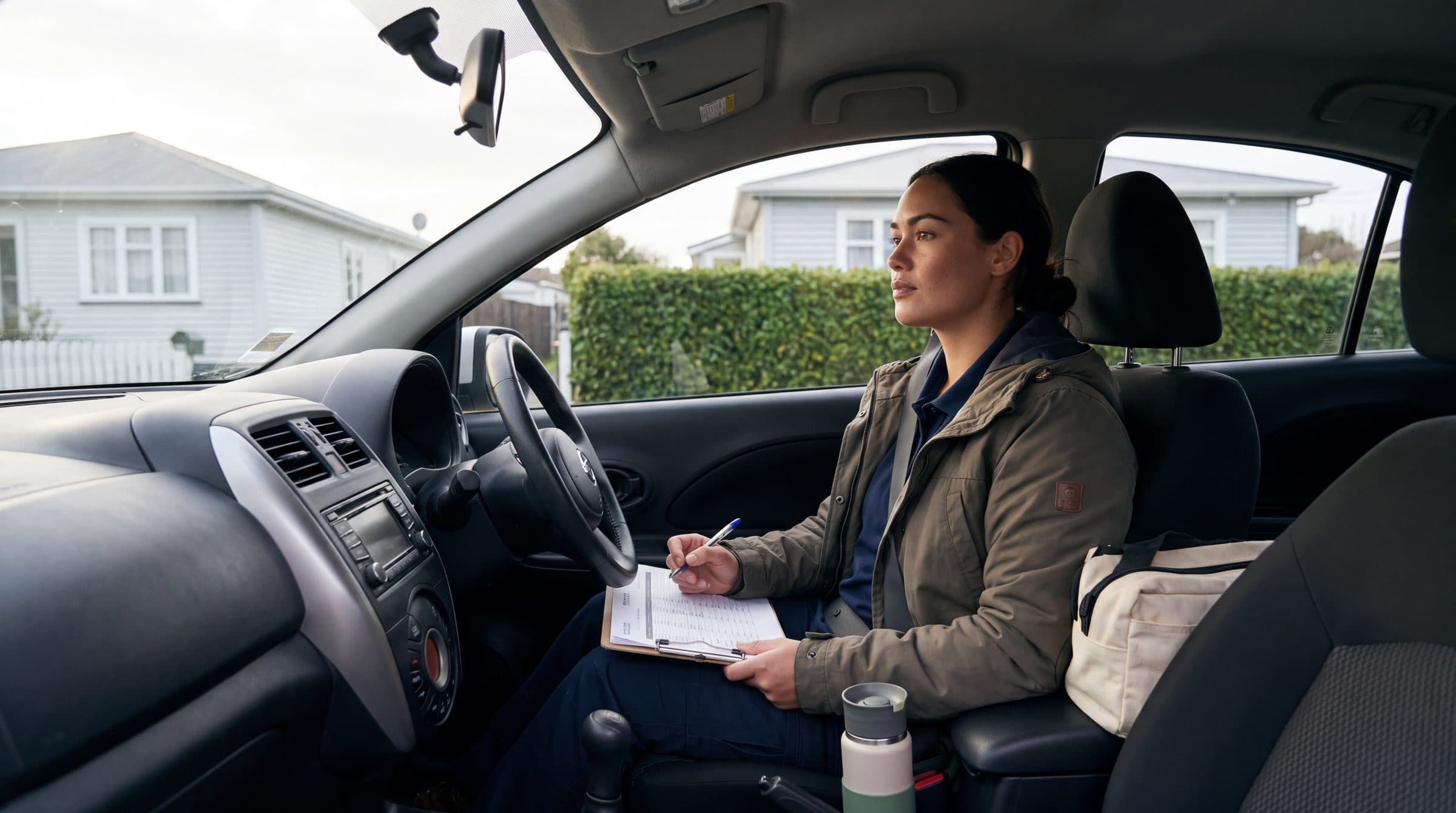 A community health worker sits in her parked car with a clipboard on her lap, pen in hand, looking out at the street