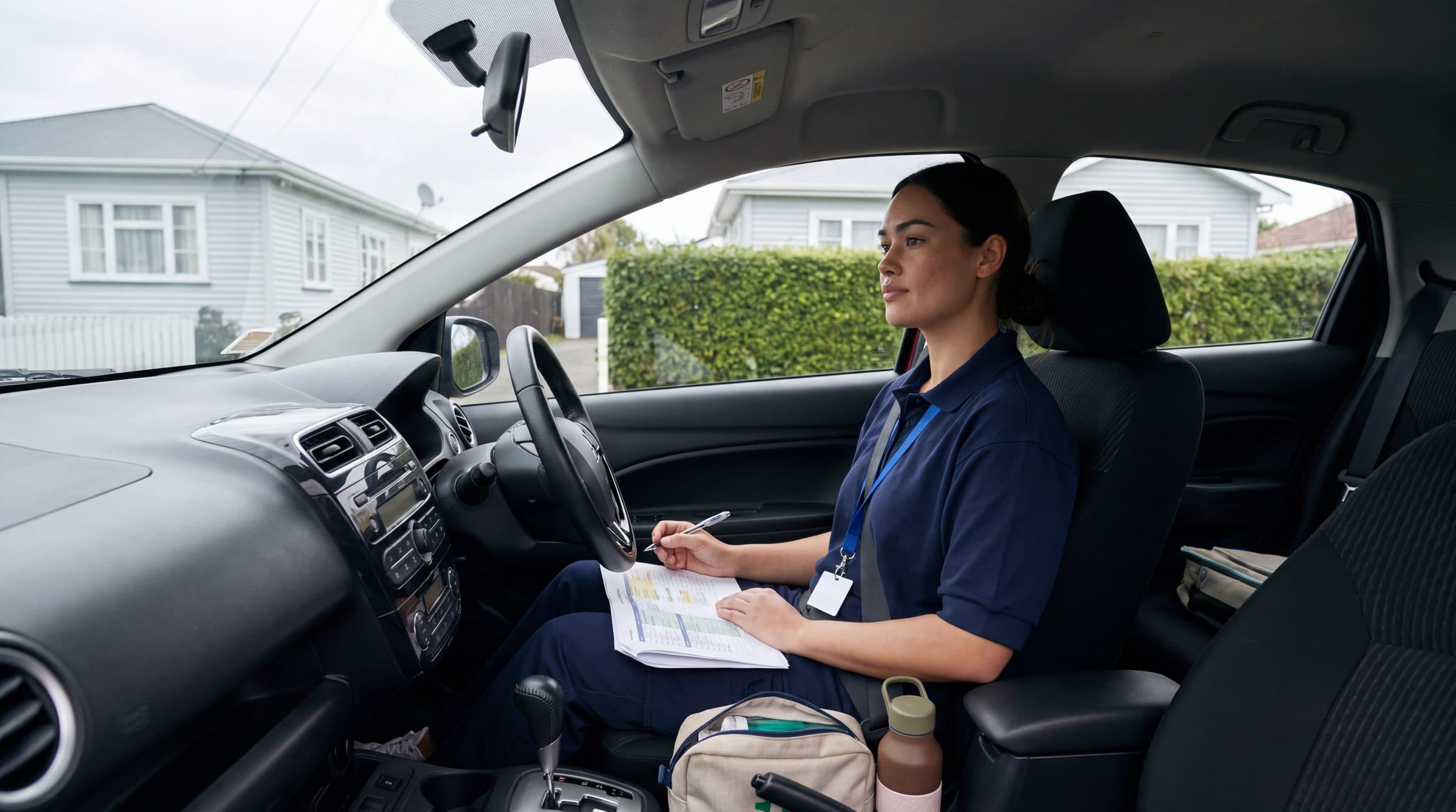A community health worker in a navy polo and lanyard sits in her parked car, writing on a printed visit schedule