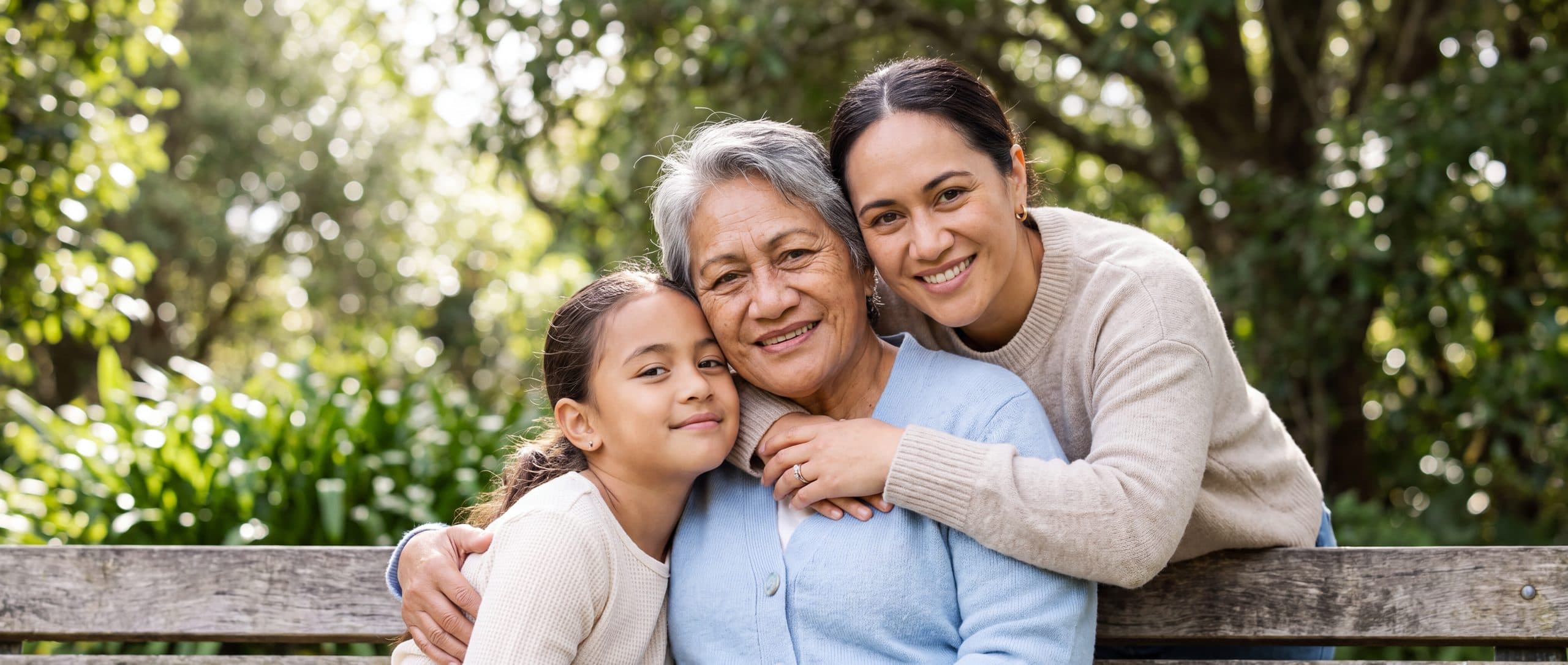 Three generations of one family sit together on a park bench under trees, the youngest leaning into her grandmother