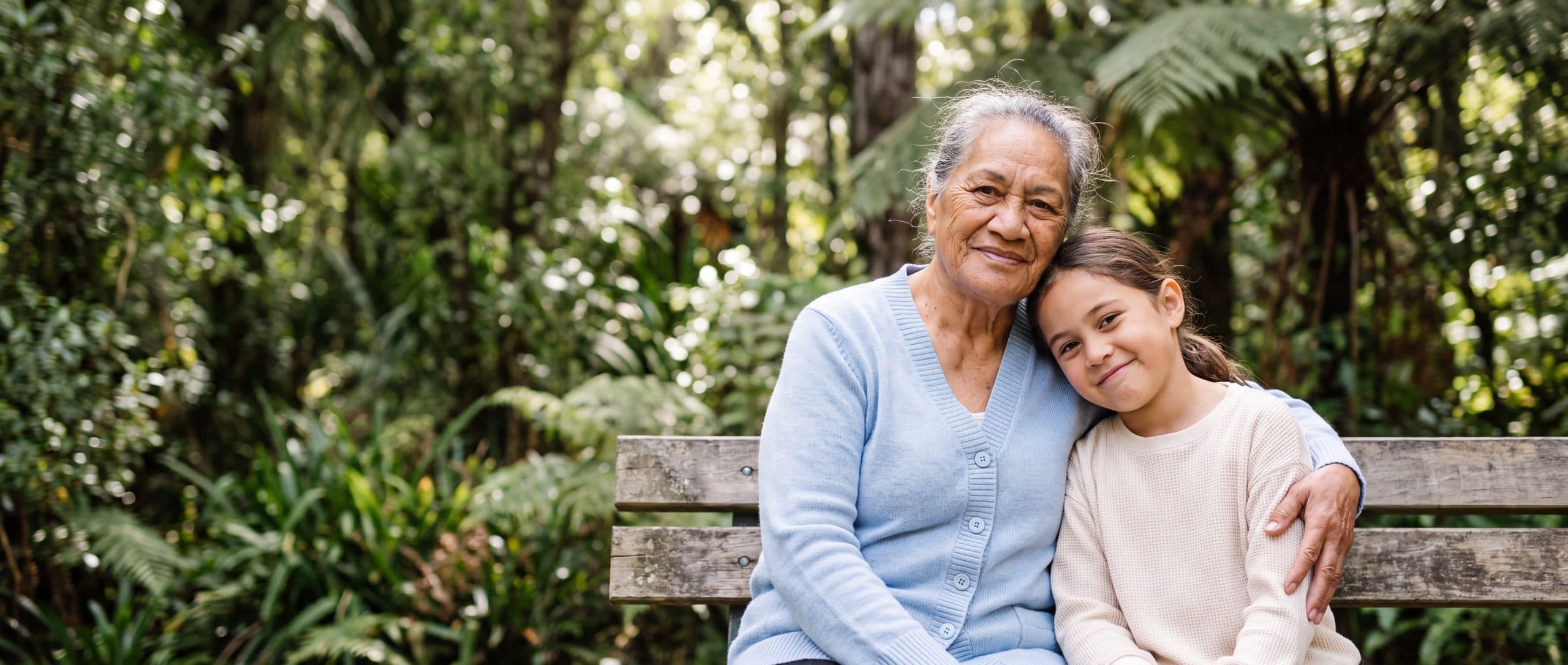 A grandmother and her granddaughter sit together on a bench in native bush, the girl resting her head on her grandmother's shoulder