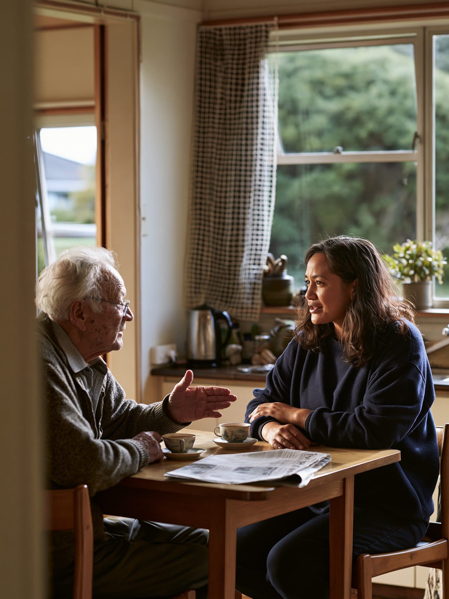 An older man talks with a visiting support worker at his kitchen table, cups of tea between them and the garden through the window