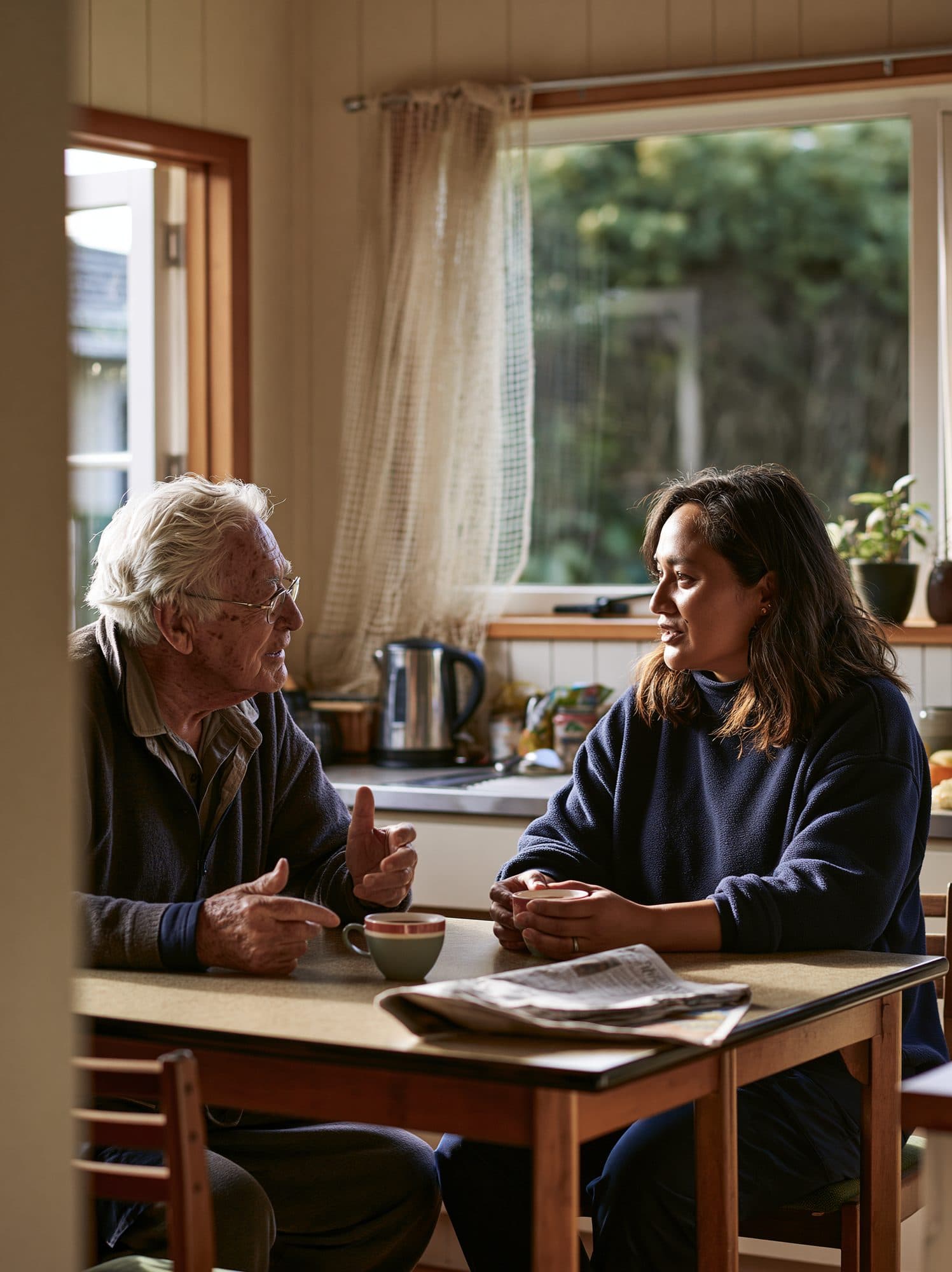 A visiting support worker listens while an older man explains something at his kitchen table, both holding cups of tea