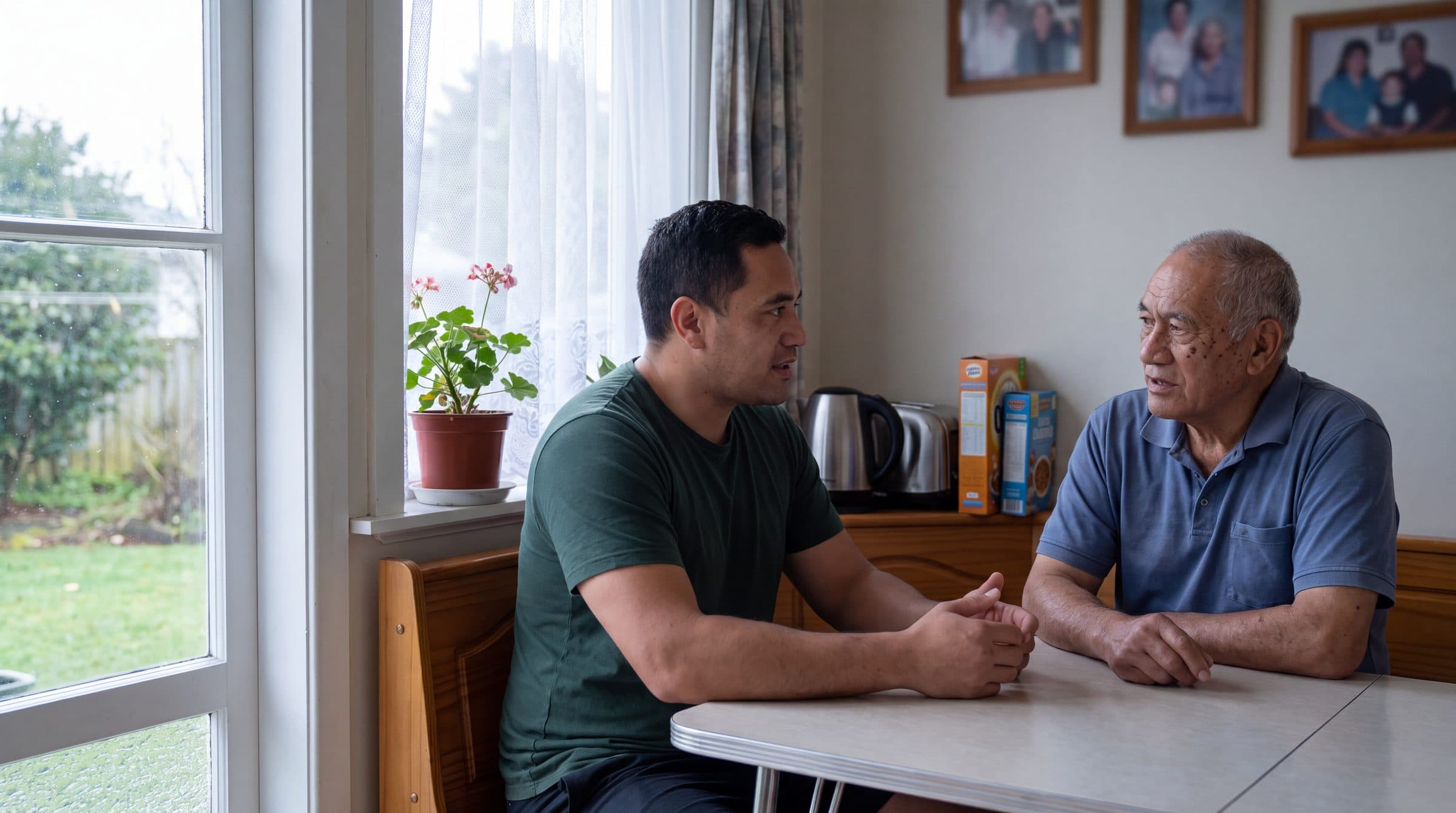 Two men talk across a kitchen table by the window, family photographs framed on the wall behind them