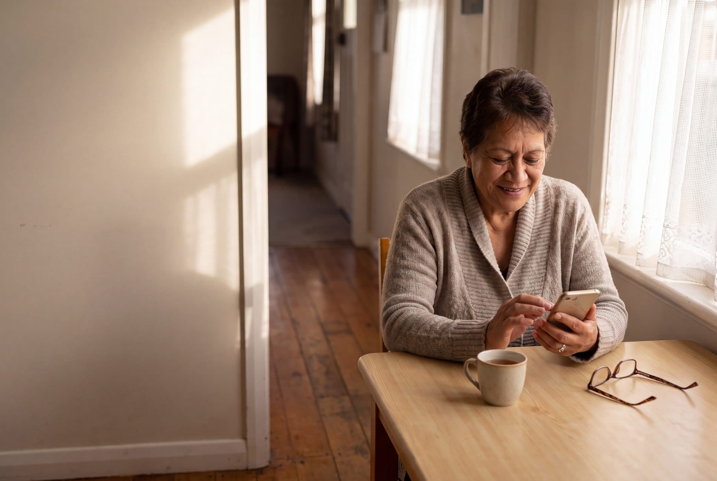 An older woman smiles at her phone at a sunlit kitchen table, a cup of tea and her reading glasses beside her