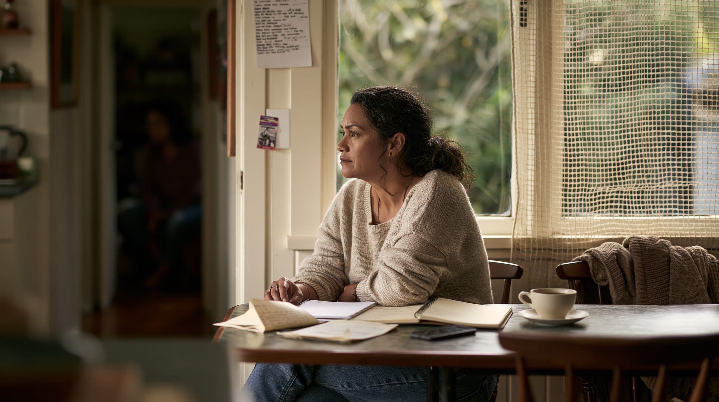 A woman sits at her kitchen table with notebooks and letters spread in front of her, looking out the window