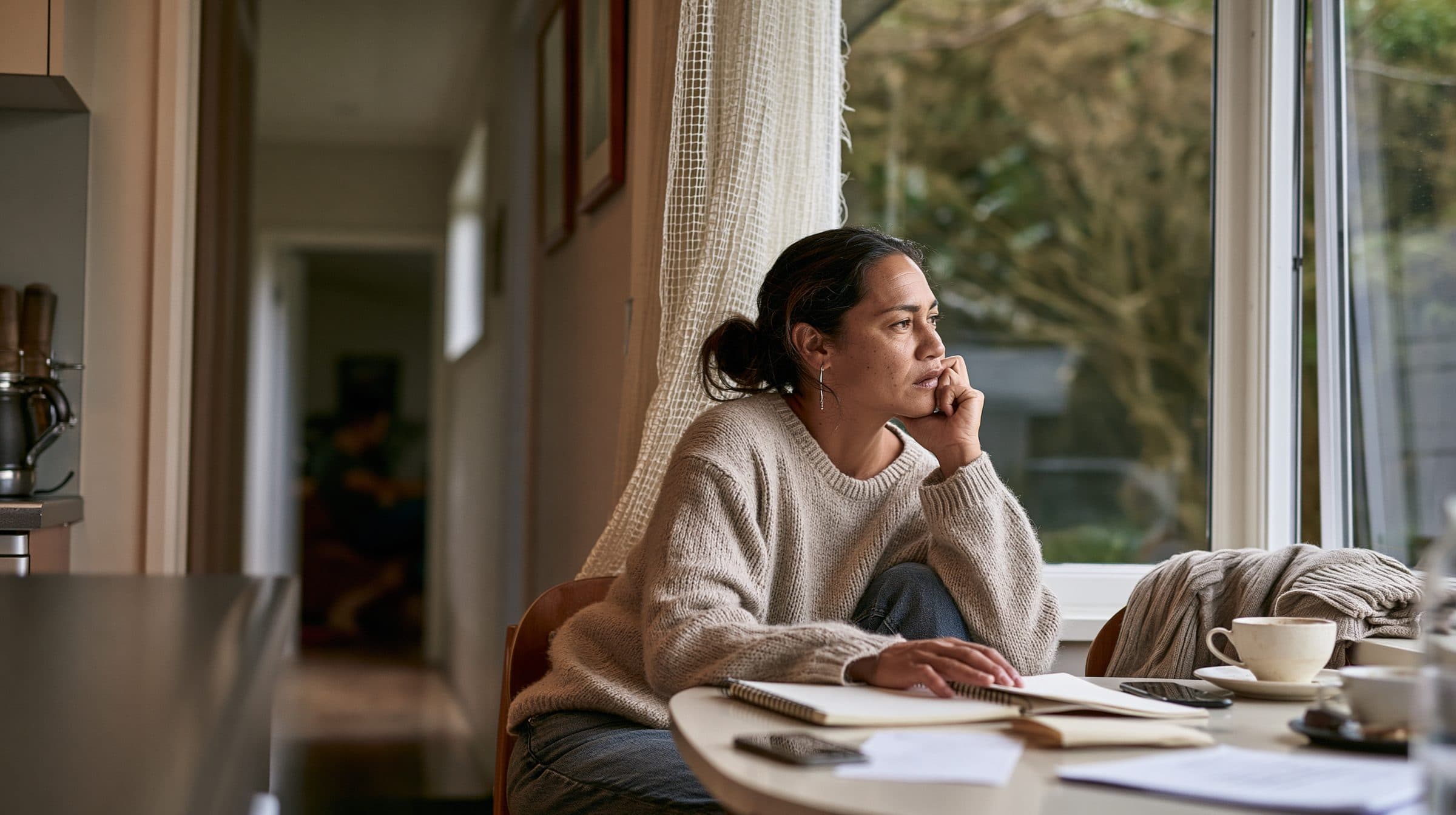 A woman rests her chin on her hand at her kitchen table, an open notebook in front of her, looking out the window